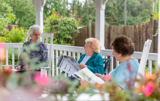 Three senior women seated outdoors in chairs surrounded by flowers. The women are talking and have open magazines in their hands.